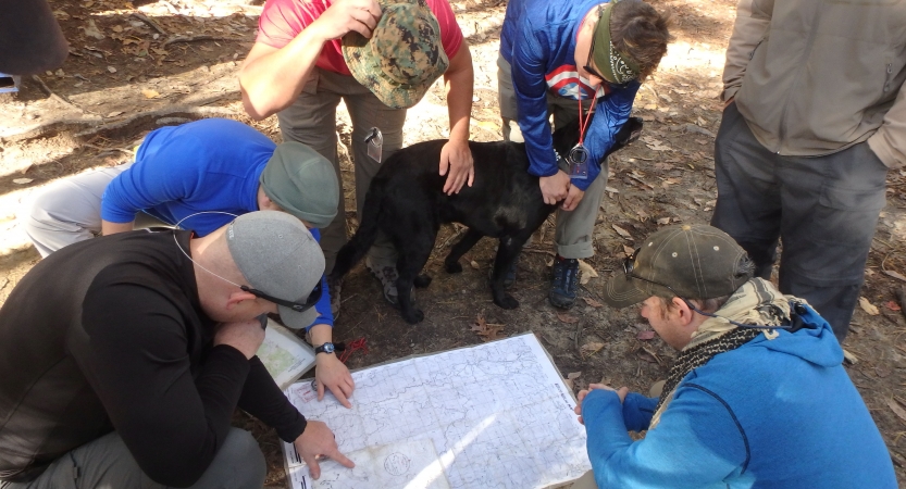 A group of people and a dog gather around a map spread out on the earth. 